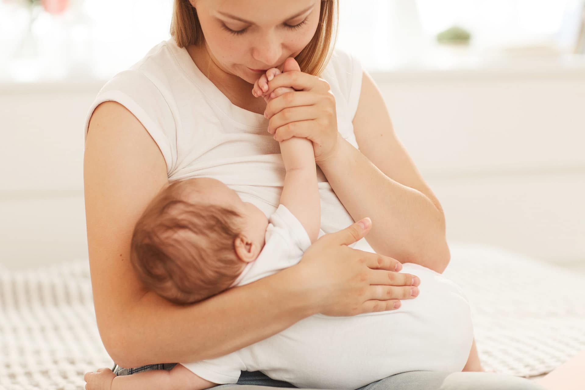 Mother smelling and kissing hand of adorable baby in bodysuit sleeping on her laps
